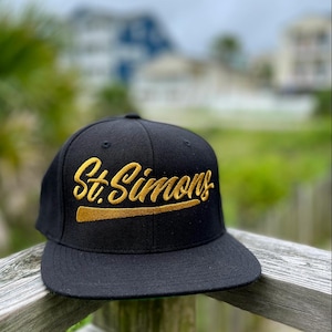 May include: A black baseball cap with the embroidered text "St. Simons" in gold script. The hat has a flat brim and a structured fit. The background is blurred, showing greenery and a hint of buildings.