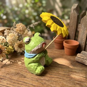 May include: A hand-carved wooden frog figurine, holding a yellow sunflower umbrella. The frog is green with a blue scarf and a surprised expression. The figurine is on a wooden surface, with small terracotta pots and a wooden fence in the background.