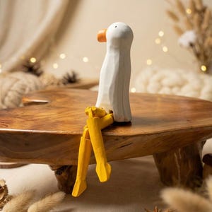 Può includere: Una statuetta di oca in legno con corpo bianco, becco arancione e zampe gialle. L'oca è seduta su un tavolo di legno. La statuetta ha un aspetto intagliato e un design stravagante.