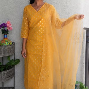 May include: A bright yellow three-piece outfit consisting of a V-neck kurta with three-quarter sleeves, matching straight-leg trousers, and a sheer organza dupatta. The kurta and trousers feature a repeating metallic gold pattern throughout.