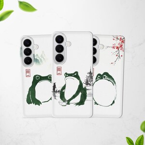 Può includere: Tre custodie bianche per telefono con un design di ispirazione giapponese. Ogni custodia presenta una diversa illustrazione di rana verde, con elementi come una pagoda, fiori di ciliegio e una barca. Le custodie hanno una finitura lucida e un ritaglio per la fotocamera.