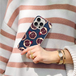 May include: A navy blue phone case featuring a pattern of pomegranate fruit slices. The case is held against a striped sweater. The pomegranates are a deep red color with a white interior and red accents.
