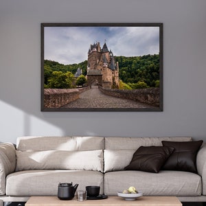 以下が含まれることがあります： 入り口に通じる石橋のある中世の城の額縁付き写真。城は木々に囲まれ、曇り空が広がっています。