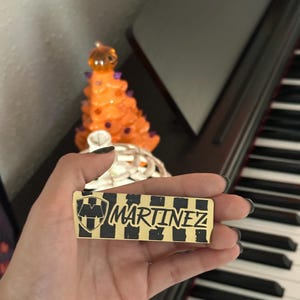 May include: A rectangular gold-colored pin with the name "MARTINEZ" in black lettering. The pin features a black and gold checkered pattern and a shield emblem. The pin is held in a hand, with a piano and decorative items in the background.