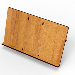 May include: A wooden book stand with a light brown finish and dark brown edges. The stand has a slanted back and a small ledge at the bottom to hold books. There are three small rectangular cutouts on the front.