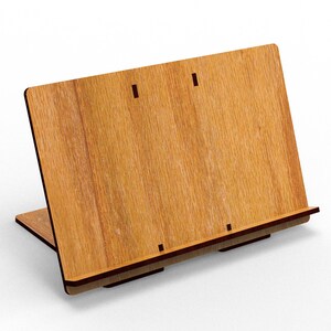 May include: A wooden book stand with a light brown finish. The stand is angled with a base and a back panel, featuring a slot for holding books. The wood grain is visible, and the stand appears to be made of layered wood.