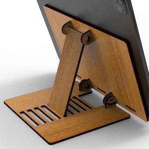 May include: A wooden tablet stand with a natural wood grain finish. The stand is designed with a base, a supporting arm, and a back panel to hold the tablet. The base has several rectangular cutouts. The tablet is black.