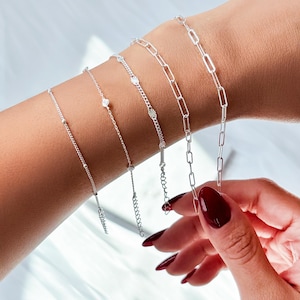 Könnte beinhalten: Eine Auswahl an silbernen Armbändern mit verschiedenen Designs, darunter filigrane Ketten mit kleinen Perlen, ein Solitär-Anhänger und ein Kettenglied-Stil. Die Armbänder werden an einem Handgelenk präsentiert und zeigen ihre unterschiedlichen Texturen und Stile.