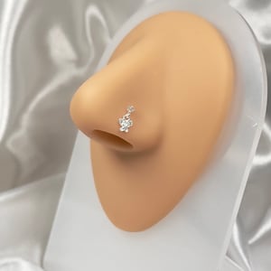 Könnte beinhalten: Ein silberner Nasenring mit einem kleinen, klaren Kristallblumen-Anhänger. Der Ring befindet sich an der Nase einer Puppe.