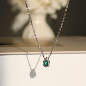 Pode incluir: Um colar de corrente de prata com um pingente em forma de lágrima que apresenta uma gema verde cercada por gemas transparentes menores.