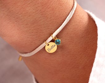 May include: A white cord bracelet with a gold charm that says "Brilla" and a small blue gemstone.