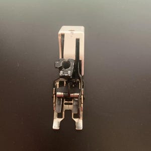 May include: A silver sewing machine foot with a black plastic attachment. The foot has a small hole in the center and a metal bar on the bottom.