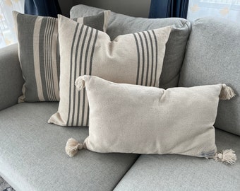 May include: A collection of decorative throw pillows in neutral tones. Two square pillows feature vertical gray stripes on a cream background. A rectangular pillow with tassels at each end rests in front. The pillows are arranged on a gray sofa.