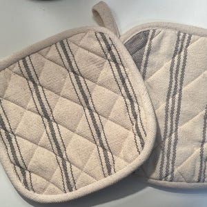 May include: Two quilted pot holders with a white background and gray stripes. The pot holders are made of a soft, textured fabric.