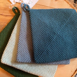 May include: A set of four square, hand-knitted cotton potholders featuring a textured garter stitch pattern. Each potholder is a different solid colour, including dark forest green, light sage green, slate blue, and deep teal, with a small loop.