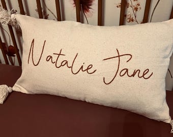 Könnte beinhalten: Ein weißes Kissen mit einem braunen gestickten Namen "Natalie Jane" auf einer burgunderfarbenen Oberfläche.
