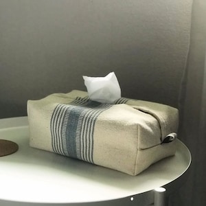 Può includere: Un copri scatola di fazzoletti bianco con strisce blu e bianche. Il copri scatola è realizzato in tessuto tessuto e ha una forma rettangolare.
