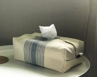 Puede incluir: Una funda para caja de pañuelos de papel blanca con rayas azules y blancas. La funda está hecha de tejido tejido y tiene una forma rectangular.
