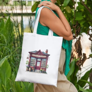 A woman carrying a white tote bag with a whimsical vintage shop illustration. The floral storefront print enhances the bag’s charm, making it perfect for eco-friendly shopping, personalized gifts, or stylish everyday accessories.