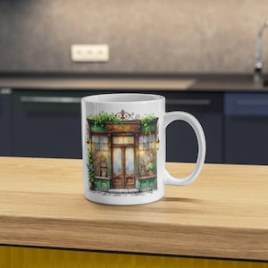 A white ceramic mug featuring a watercolor vintage shop illustration. The elegant storefront print adds a nostalgic touch, perfect for coffee lovers, kitchen decor, or as a thoughtful personalized gift for any occasion.
