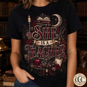 Pode incluir: Uma t-shirt preta com um design gráfico contendo o texto "SHE IS A TEACHER" numa fonte decorativa. A ilustração inclui um livro aberto, uma lua crescente, uma vela acesa, rosas vermelhas, uma maçã vermelha, óculos, uma caneta e uma prancheta.
