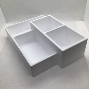Op de afbeelding: Een witte organizer met meerdere compartimenten met drie secties van verschillende afmetingen. De organizer is gemaakt van een solide, ondoorzichtig materiaal en is ontworpen om kleine items in op te bergen. De compartimenten zijn rechthoekig en hebben een strak, minimalistisch ontwerp.