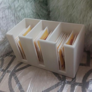 May include: A white plastic tea bag organizer with four compartments, each filled with tea bags. The organizer is rectangular with a simple, clean design, perfect for kitchen or office use.