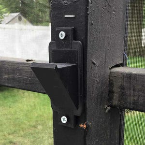May include: A black gate latch attached to a wooden fence post. The latch is made of a dark material and is secured with screws. The fence has a black frame and a mesh screen. The background shows a grassy area and a house.