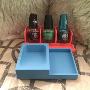 May include: A pink and blue nail polish organizer holding three bottles of nail polish. The nail polish bottles are various colors, including red, green, and teal. The organizer has a blue tray for additional items.