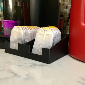 Puede incluir: Un organizador de bolsitas de té de plástico negro con dos compartimentos separados, cada uno lleno de una fila de bolsitas de té blancas con etiquetas de papel amarillo. El organizador tiene un perfil frontal bajo y lados más altos.