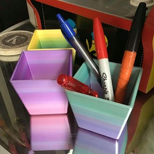 Op de afbeelding: Vier vierkante bakjes met open bovenkant in een kleurengradient van geel naar paars. Elk bakje bevat pennen en stiften, waaronder een rode Sharpie. De bakjes staan op een reflecterend oppervlak.