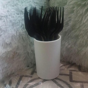 May include: A collection of black plastic forks are neatly arranged inside a white cylindrical container. The forks are made of a matte black plastic and are standing upright. The container is smooth and opaque, with a simple, minimalist design.
