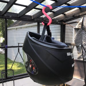 May include: A black motorcycle helmet is suspended from a pink S-shaped hook. The helmet has a clear visor and a black strap. The hook is attached to a black structure with a clear roof. A blue strap is also visible.