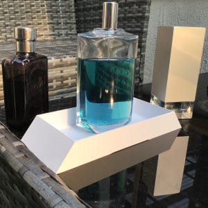 May include: A clear glass perfume bottle filled with bright blue liquid, a dark purple glass bottle with a silver cap, and a tall white rectangular box, all arranged together on a white tray with angled sides and a dark base.