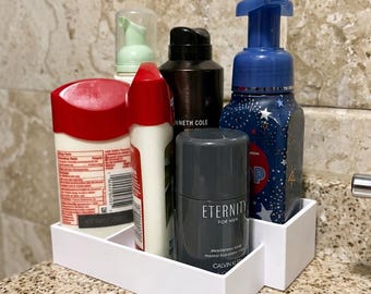 Puede incluir: Una bandeja organizadora de plástico blanco que contiene varios artículos de cuidado personal, incluyendo una botella blanca con tapa roja, una botella más pequeña con tapa roja, una botella negra Kenneth Cole, un desodorante gris Calvin Klein Eternity y un dispensador de jabón espumoso azul.