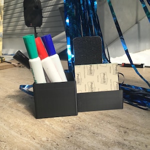 May include: A black desk organizer with two compartments. One side holds four markers in green, red, blue, and black. The other side holds a black rectangular eraser and a stack of square Post-it notes with the brand name printed on each sheet.