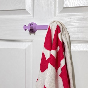 May include: A light purple, horizontal wall-mounted towel hook with a circular base and a T-shaped end, holding a white and bright pink chevron-patterned towel. The hook is secured with a visible screw and extends outward from the flat surface.