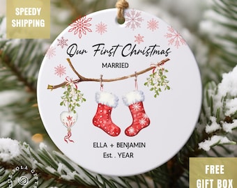 May include: A round Christmas ornament with the text "Our First Christmas Married". The ornament features red stockings, snowflakes, and mistletoe. The names "Ella + Benjamin" and the year are also included. The ornament is hanging on a Christmas tree.