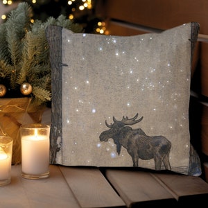 Puede incluir: Una almohada beige con una silueta de alce marrón y puntos blancos sobre un fondo de bosque nevado.