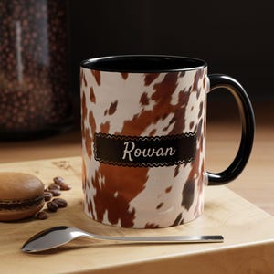 May include: A black and white coffee mug with a brown cowhide pattern and the name "Rowan" printed on a black banner. The mug has a black handle.