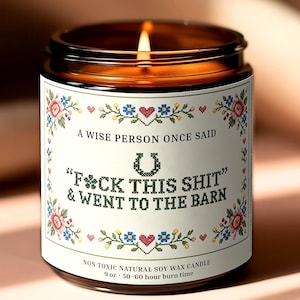 May include: A brown glass jar candle with a white label featuring cross-stitch style floral borders and the text: A WISE PERSON ONCE SAID F*CK THIS SHIT & WENT TO THE BARN. The label includes a horseshoe icon and states 9 oz.