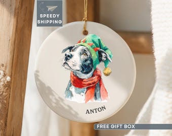 May include: A round white ceramic ornament hanging from a gold string. The ornament features a watercolor illustration of a black and white dog wearing a green elf hat and a red scarf. The name ANTON is printed in black text below the dog.