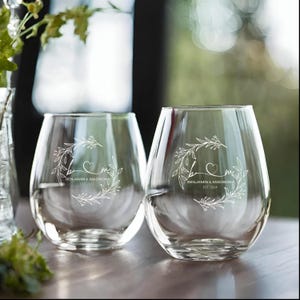 Op de afbeelding: Twee heldere, stamlloze wijnglazen met een decoratief bloemenkransontwerp. De glazen zijn gegraveerd met de initialen "b & m", een hart, de namen "Benjamin & Madison" en het jaar "EST 2024".