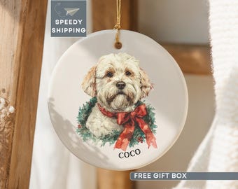 May include: A round white ceramic ornament featuring a watercolor illustration of a cream-colored dog wearing a red bow and a green wreath around its neck. The name COCO is printed in black text below the dog. It hangs from a gold string.