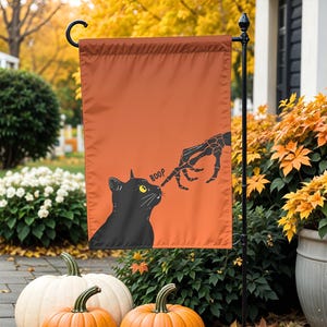 Może przedstawiać: Pomarańczowa flaga ogrodowa na Halloween z czarnym kotem i szkieletową dłonią. Kot jest dotykany po nosie przez szkieletową dłoń, z napisem "BOOP" powyżej. Flaga jest wystawiona na zewnątrz z dyniami i jesiennymi liśćmi.