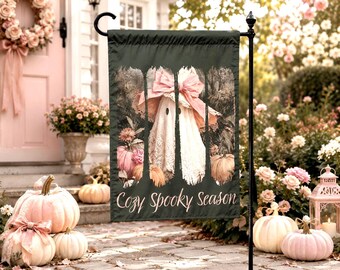 May include: A dark green garden flag featuring a central illustration of a ghost wearing a large pink bow, surrounded by pumpkins and flowers. The text "Cozy Spooky Season" is printed in a white script font at the bottom of the rectangular flag.