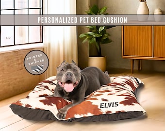 May include: A rectangular pet bed cushion featuring a brown and white cowhide print pattern with the name ELVIS printed in black. A gray dog is lying on the cushion. The text PERSONALIZED PET BED CUSHION is visible at the top of the image.