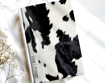May include: A hardcover notebook featuring a black and white cow print pattern across the front cover. The text "DANIELLE'S NOTES" is printed in a vertical, sans-serif black font along the left side of the cover near the spine.