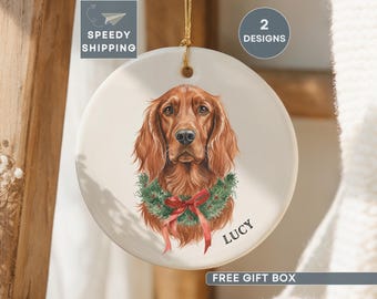 May include: A round, cream-colored ceramic ornament featuring a detailed illustration of a red-haired dog with a green wreath and red bow. The name "LUCY" is printed below the dog. The ornament has a gold string for hanging.