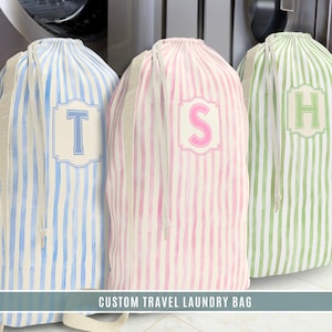 Puede incluir: Tres bolsas de lavandería de viaje personalizadas con rayas verticales azules, rosas y verdes. Cada bolsa presenta una inicial monogramada en un marco decorativo: "T", "S" y "H". Las bolsas tienen cierres de cordón y están hechas de tela ligera.